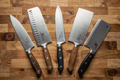 Collection de couteaux de cuisine professionnels disposés en éventail sur une planche en bois, incluant couteau de chef, couteau à pain, couteau d'office et santoku